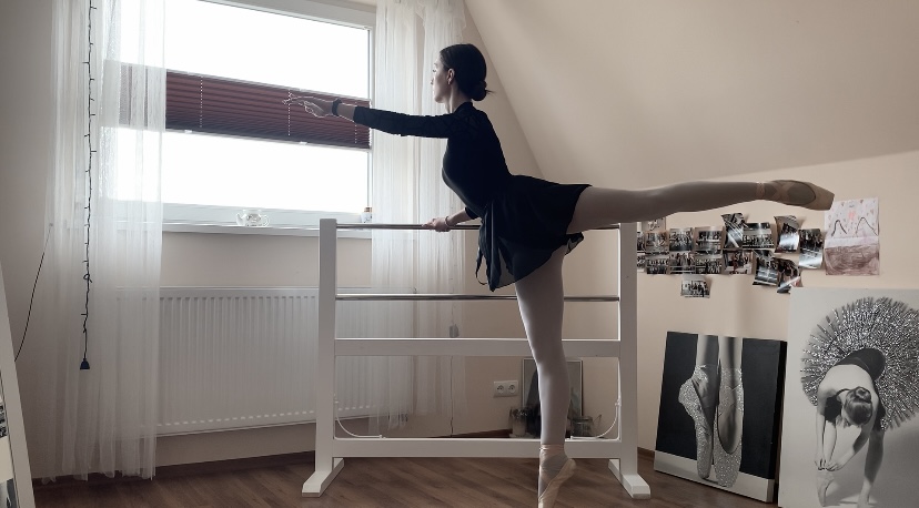 Kasia w swoim pokoju w stroju baletnicy wykonuje ćwiczenia przy drążku baletowym na pointach: stoi na palcach jednej nogi, druga prosta uniesiona, ręka wyciągnięta przed siebie