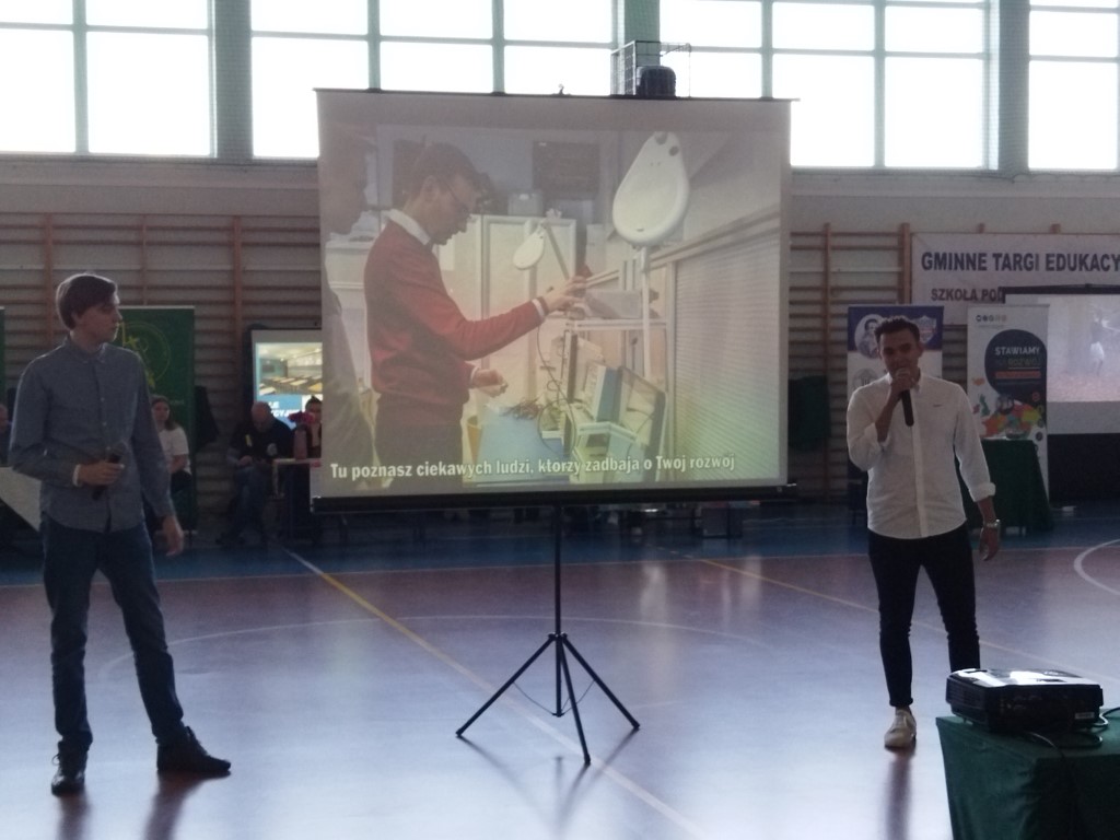 Na środku sali ekran na stojaku, wyświetlane kadry filmu. Z prawej strony uczeń w białej koszuli z mikrofonem. Po lewej stronie uczeń w niebieskiej koszuli.