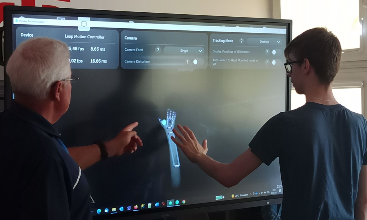 Ekran monitora z wyświetloną ręką. Przed monitorem nauczyciel i uczeń, który dotyka ekranu.