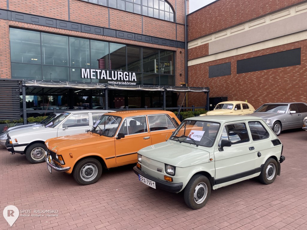 Wystawa starych samochodów. Na pierwszym planie Fiat 126p, za nim Fiat 125p., dalej Polonez. Samochody stoją przed Wejściem do nowoczesnego budynku. Napis na szkle "Metalurgia".