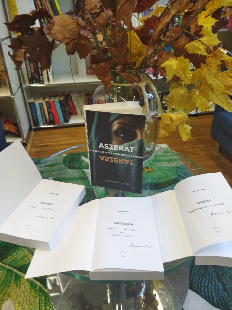 Na szklanym stoliku wyłożone książki z dedykacjami. Za otwartmi książkami stoi pozycja "Asherad", Z tyłu stolika wazon z jesienną kompozycją.