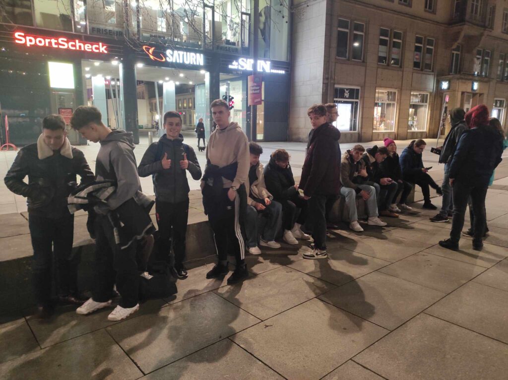 Grupa uczniów siedzi na murku przed sklepem.