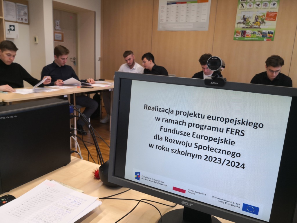 Na pierwszym planie widać monitor na którym wyświetlany jest slajd z informacją o realizacji projektu europejskiego w ramach programu FERS. W tle uczniowie siedząc w ławkach wypisują dokumenty projektowe.